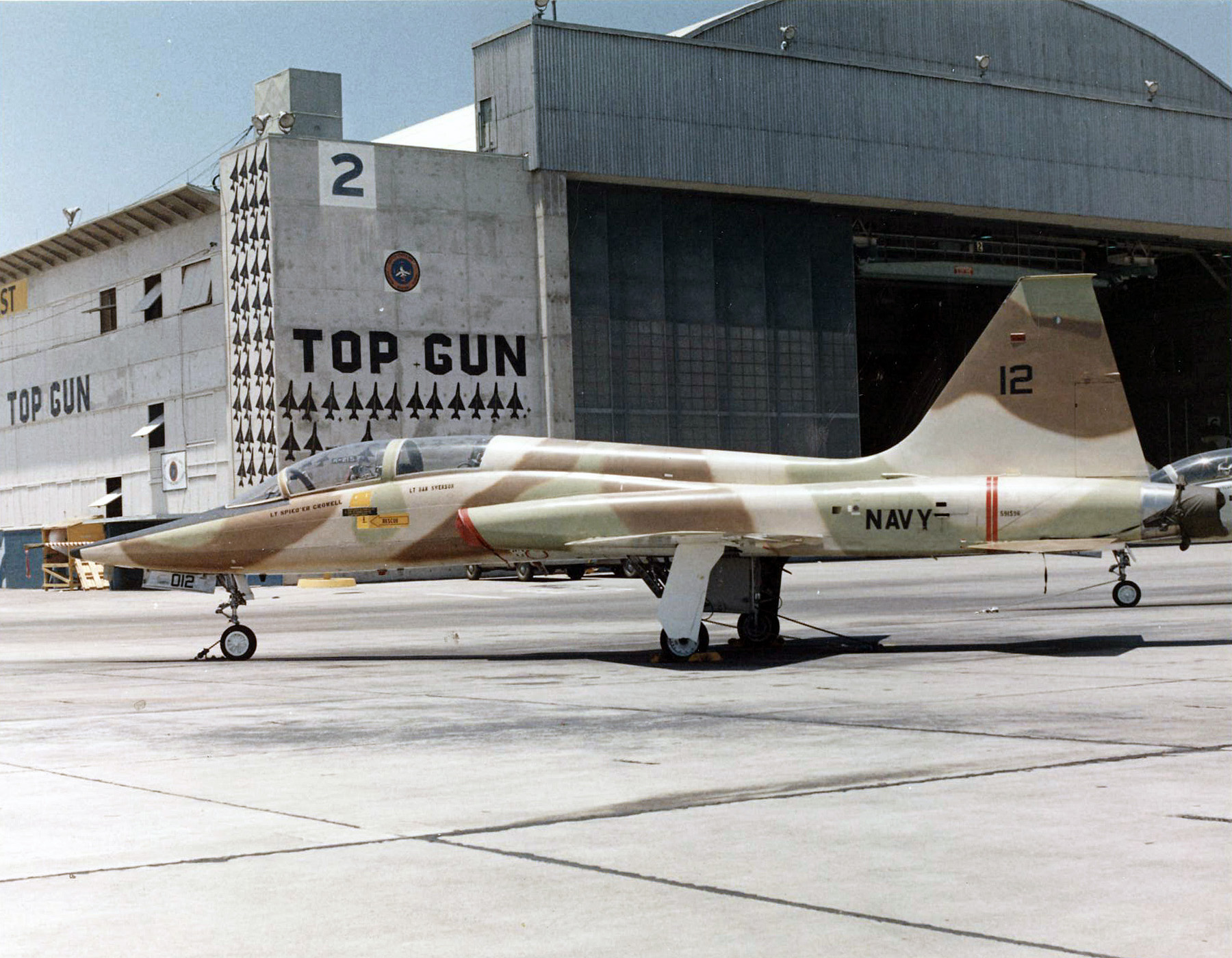 A T-38A Talon assigned to the Navy Fighter Weapons School pictured in front of the TOPGUN hangar on board Naval Air Station (NAS) Miramar, California, in 1974. The Talon was one of the aircraft types employed as an aggressor for training at the school. Note the silhouettes of MiG aircraft shot down during the Vietnam War painted on the hangar exterior. (Robert L. Lawson Photograph Collection, National Naval Aviation Museum)